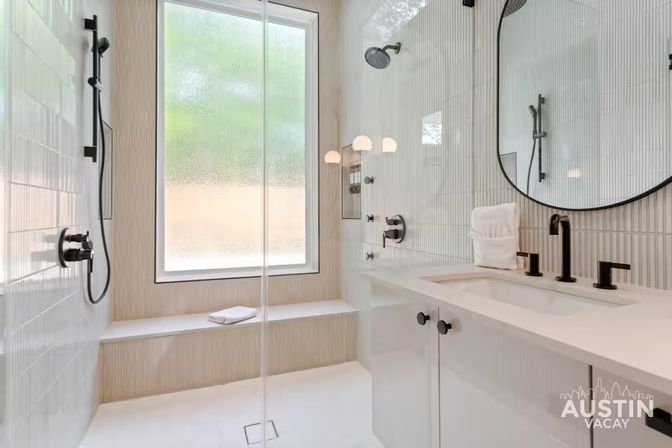 Bright, spa-like modern bathroom with glass walk-in shower, frosted window and built-in bench, white vertical tiles, round mirror and matte black fixtures over a white vanity.