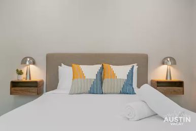 Bright modern guest bedroom with gray upholstered headboard, crisp white bedding, two yellow-and-blue geometric throw pillows, rolled white towel at foot, and floating wooden nightstands with silver dome lamps and a small potted plant.