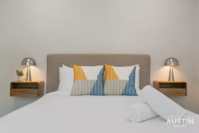 Bright modern guest bedroom with gray upholstered headboard, crisp white bedding, two yellow-and-blue geometric throw pillows, rolled white towel at foot, and floating wooden nightstands with silver dome lamps and a small potted plant.