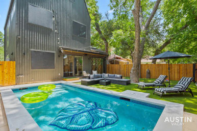 Modern Austin backyard with rectangular swimming pool and neon float rings, outdoor sectional sofa, striped chaise lounges and umbrella beneath mature oak trees beside a black two-story house and wooden fence.