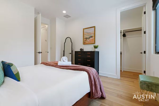 Bright, modern vacation rental bedroom with white walls and hardwood floors, queen bed with white linens and mauve throw, black dresser and full-length mirror, decorative landscape painting and open walk-in closet.