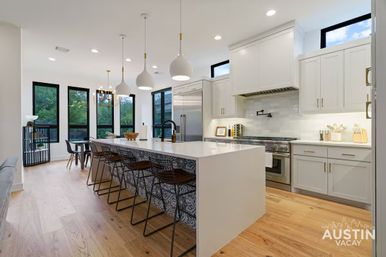 Bright modern Austin, TX kitchen with a long white waterfall island, patterned tile island base, black wire bar stools, pendant lights, stainless appliances, black-framed windows and light hardwood floors, perfect for entertaining.