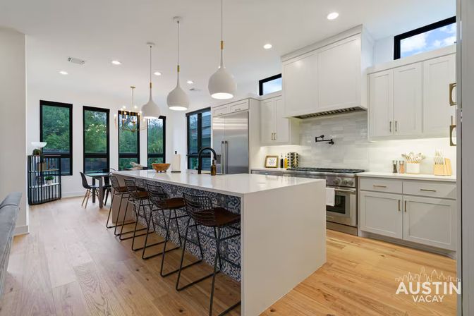 Bright modern Austin, TX kitchen with a long white waterfall island, patterned tile island base, black wire bar stools, pendant lights, stainless appliances, black-framed windows and light hardwood floors, perfect for entertaining.