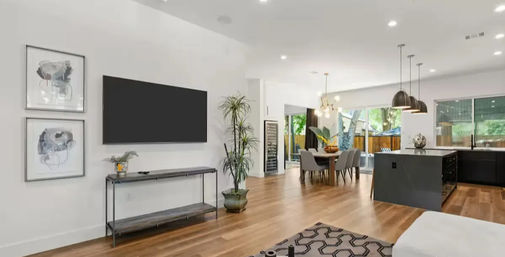 Modern open-concept living room and kitchen with wall-mounted TV, console table, abstract wall art, tall potted plant, warm hardwood floors, dining table under a chandelier, black island with pendant lights, and sliding glass doors to a fenced backyard.