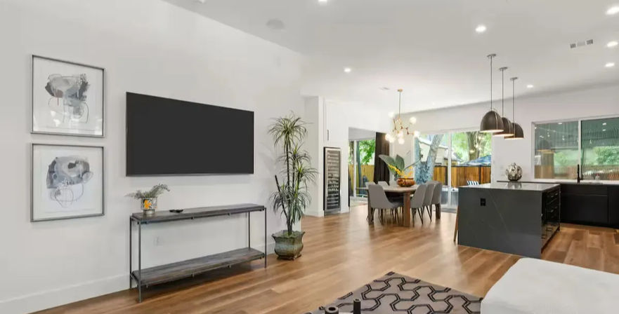 Modern open-concept living room and kitchen with wall-mounted TV, console table, abstract wall art, tall potted plant, warm hardwood floors, dining table under a chandelier, black island with pendant lights, and sliding glass doors to a fenced backyard.