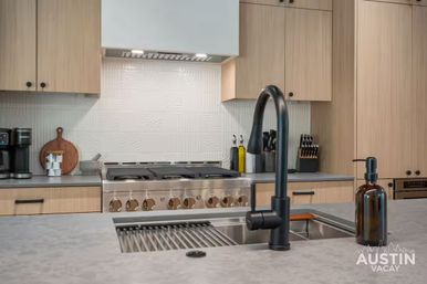Sleek Austin kitchen island with matte-black gooseneck faucet, amber soap dispenser, stainless gas range, light wood cabinets, knife block and coffee maker.