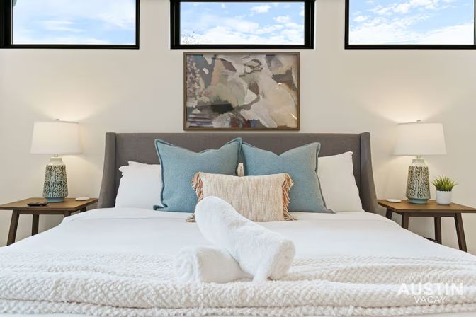 Bright Austin vacation-rental bedroom with crisp white linens, rolled towels on the bed, teal accent pillows, bedside lamps, abstract wall art and high transom windows