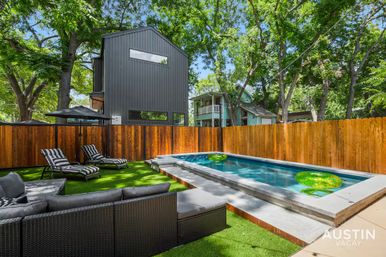 Austin backyard with modern black metal-clad house, wooden privacy fence, rectangular plunge pool with green float rings, artificial turf, striped chaise lounges and wicker outdoor sofa under tall trees.