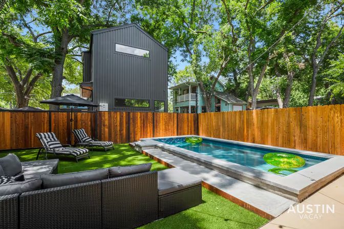 Austin backyard with modern black metal-clad house, wooden privacy fence, rectangular plunge pool with green float rings, artificial turf, striped chaise lounges and wicker outdoor sofa under tall trees.