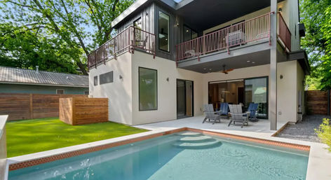 Modern two-story home backyard oasis with rectangular pool, covered patio seating, upper balcony, artificial lawn and wood fence under leafy trees.