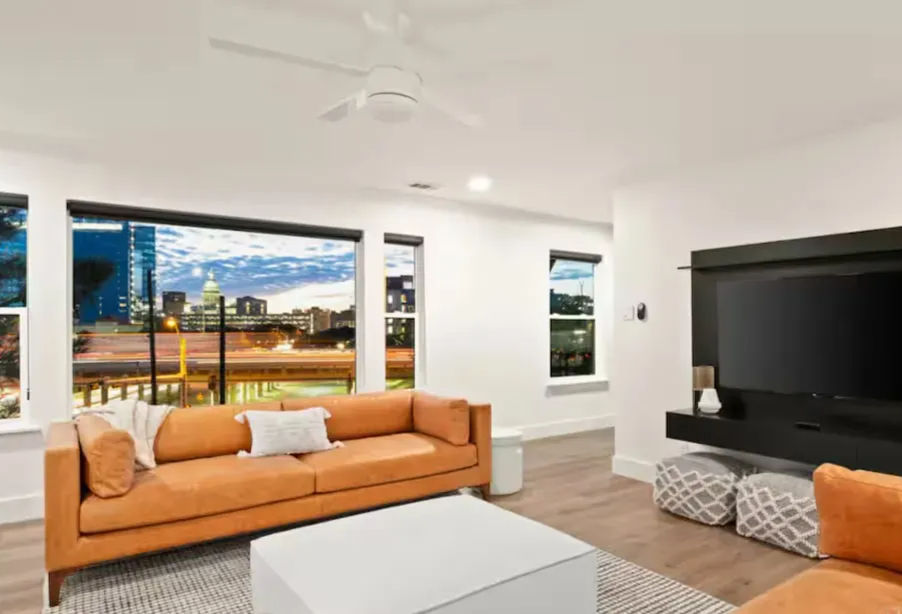 Modern living room with orange sofas, white walls, ceiling fan, wall-mounted TV and large windows showcasing a city skyline at dusk.