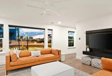 Modern living room with orange sofas, white walls, ceiling fan, wall-mounted TV and large windows showcasing a city skyline at dusk.