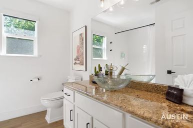 Sunlit modern bathroom with speckled granite countertop, textured glass vessel sink and brass faucet, white cabinets, large mirror, toilet, framed art and a leafy window view.