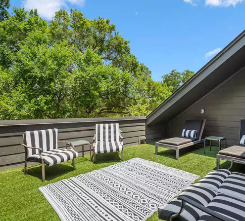 Sunny tree-lined rooftop patio with artificial turf, black-and-white striped lounge chairs and chaise lounges, patterned outdoor rug and blue sky.