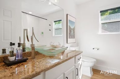 Bright modern bathroom with granite countertop, green-tinted glass vessel sink and sleek faucet, white cabinets, large mirror, toilet, wood-look floor and sunny window