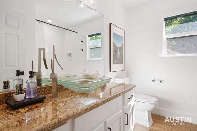 Bright modern bathroom with granite countertop, green-tinted glass vessel sink and sleek faucet, white cabinets, large mirror, toilet, wood-look floor and sunny window