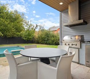 Covered poolside patio with a round table and four white chairs, modern stainless-steel built-in grill and vent hood, and a grassy tree-lined backyard under a blue sky