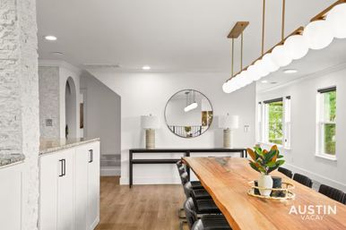 Bright Austin dining room with long live-edge wooden table, black leather chairs, row of globe pendant lights, round wall mirror, potted centerpiece and hardwood floors