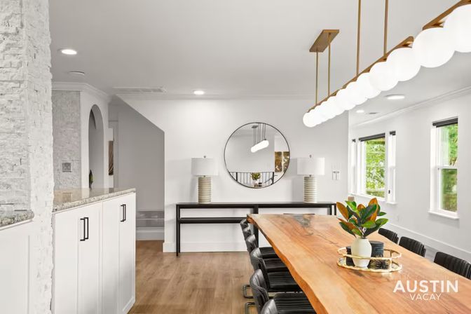 Bright Austin dining room with long live-edge wooden table, black leather chairs, row of globe pendant lights, round wall mirror, potted centerpiece and hardwood floors