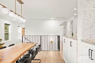 Bright open-concept dining area with a long live-edge wood table, black leather chairs, globe pendant lights, white cabinets and stacked stone accent wall by a black metal stair railing