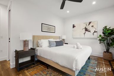 Bright, stylish Austin vacation rental bedroom with king bed dressed in white linens, abstract wall art, bedside lamps, potted plant and a colorful area rug.