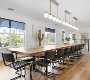 Bright modern meeting room with a long live-edge wooden conference table, row of black cantilever chairs, globe pendant lights, large windows with urban greenery views, and warm hardwood floors.
