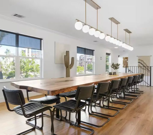 Bright modern meeting room with a long live-edge wooden conference table, row of black cantilever chairs, globe pendant lights, large windows with urban greenery views, and warm hardwood floors.