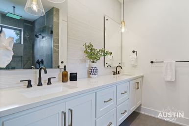 Bright spa-like modern bathroom with white double vanity, black matte faucets, large mirror, glass pendant lights, patterned vase with greenery, and a tiled walk-in shower.