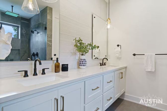 Bright spa-like modern bathroom with white double vanity, black matte faucets, large mirror, glass pendant lights, patterned vase with greenery, and a tiled walk-in shower.
