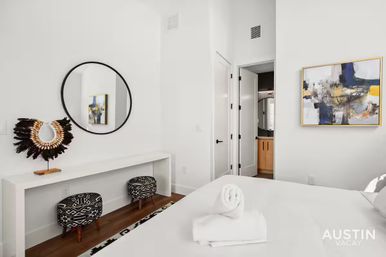 Bright, minimalist Austin apartment bedroom with white walls, round black mirror, feathered decorative collar on a console, patterned stools, abstract wall art, and rolled towels on the bed.