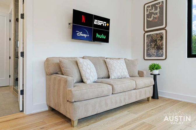 Cozy modern rental living room with a beige three-seat sofa and patterned throw pillows, wall-mounted TV showing Netflix, ESPN+, Disney+ and Hulu logos, framed art, small potted plant on a black side table, and light wood floors.