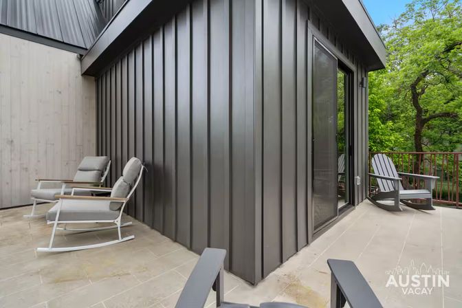 Modern elevated balcony with dark vertical metal siding, two cushioned rocking loungers and Adirondack chairs overlooking leafy trees — outdoor patio in Austin, Texas.