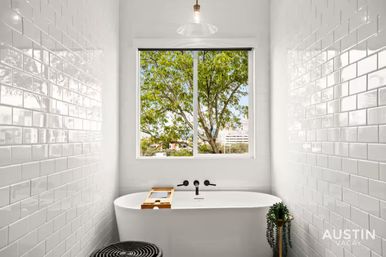 Bright modern bathroom with glossy white subway-tiled walls, freestanding bathtub beneath a window framing a leafy tree, wooden bath caddy and small potted plant.