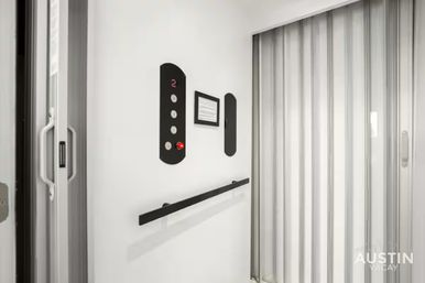 Sleek white elevator interior with folding accordion door, black handrail and vertical control panel showing illuminated '2' and a red emergency button.