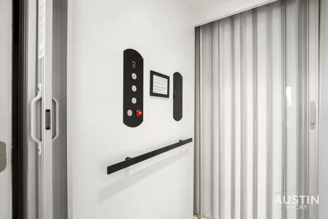 Sleek white elevator interior with folding accordion door, black handrail and vertical control panel showing illuminated '2' and a red emergency button.