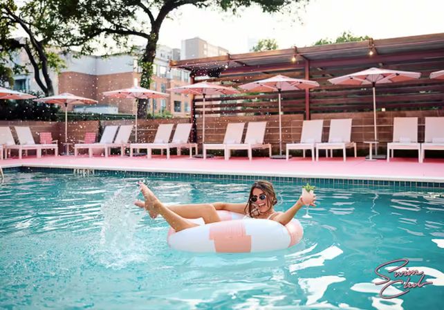 Person lounging on a pink-and-white inflatable donut splashing in a turquoise urban pool, holding a cocktail with pink deck, lounge chairs and umbrellas in the background.