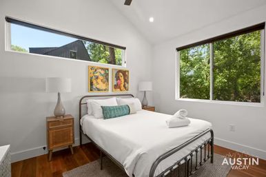 Bright Austin vacation-rental bedroom with metal bed frame, crisp white linens and teal bolster, mid-century wood nightstands with glass lamps, colorful artwork above the bed and large windows overlooking leafy trees.