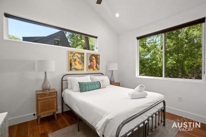 Bright Austin vacation-rental bedroom with metal bed frame, crisp white linens and teal bolster, mid-century wood nightstands with glass lamps, colorful artwork above the bed and large windows overlooking leafy trees.