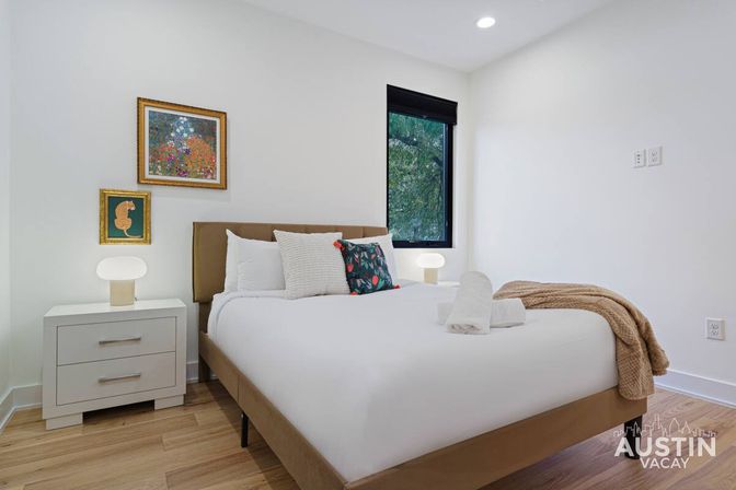 Bright minimalist Austin bedroom with plush bed in white linens, colorful accent pillows, modern nightstands and lamps, framed art, and a tree‑lined window.