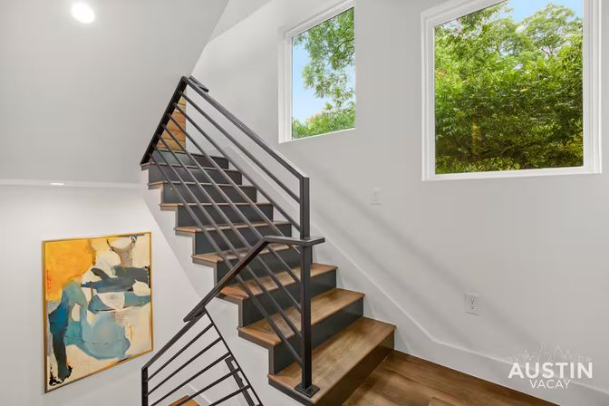 Bright modern interior staircase with sleek black metal railing and warm wood treads, two windows framing green trees, and a bold abstract painting on the wall.