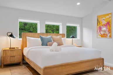 Austin vacation rental bedroom with wooden platform bed, crisp white linens and rolled towel, three windows framing green trees, rattan nightstands with black lamps and colorful horse print