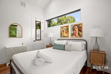 Bright modern bedroom in Austin, Texas with crisp white linens on a queen bed, colorful wall art, narrow high window showing green trees, wooden nightstands and glass lamps.