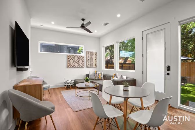 Bright modern living room in Austin with a round dining table and white chairs, gray sectional sofa, wall-mounted TV, wood floors, ceiling fan, and glass door to a leafy backyard.