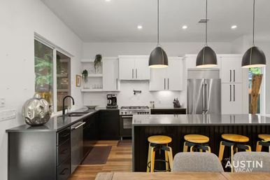 Contemporary open kitchen with black lower cabinets and white uppers, stainless fridge and range, long island with four yellow bar stools, three pendant lights and a window showing greenery.