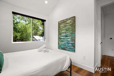 Bright, cozy bedroom with crisp white linens, large window framing leafy trees, teal abstract painting on a white wall and warm hardwood floors — Austin, TX vacation rental.