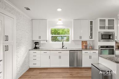Bright modern white kitchen with shaker cabinets, granite countertops, stainless steel appliances, sink under a window with green trees and hardwood floors.