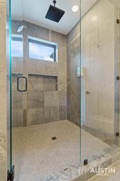 Modern gray tiled walk-in shower with frameless glass door, ceiling rain showerhead, recessed niche and wall-mounted soap dispenser under a small high window in Austin, TX bathroom
