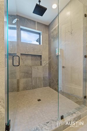 Modern gray tiled walk-in shower with frameless glass door, ceiling rain showerhead, recessed niche and wall-mounted soap dispenser under a small high window in Austin, TX bathroom