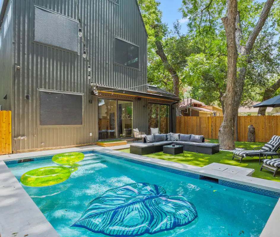 Modern two-story gray house backyard with rectangular swimming pool, neon green inner-tube floats and a blue leaf-shaped float, outdoor sectional sofa and lounge chairs on artificial turf, wooden privacy fence and large shade trees