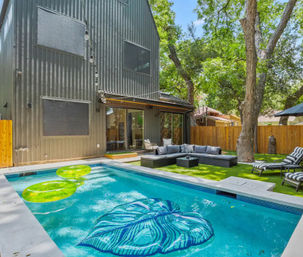 Modern two-story gray house backyard with rectangular swimming pool, neon green inner-tube floats and a blue leaf-shaped float, outdoor sectional sofa and lounge chairs on artificial turf, wooden privacy fence and large shade trees